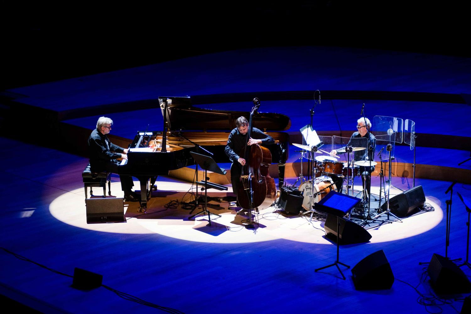 Trio Andrzeja Jagodzińskiego, pianista siedzi przy czarnym fortepianie, kontrabasista stoi z instrumentem, perkusista siedzi za składem instrumentów