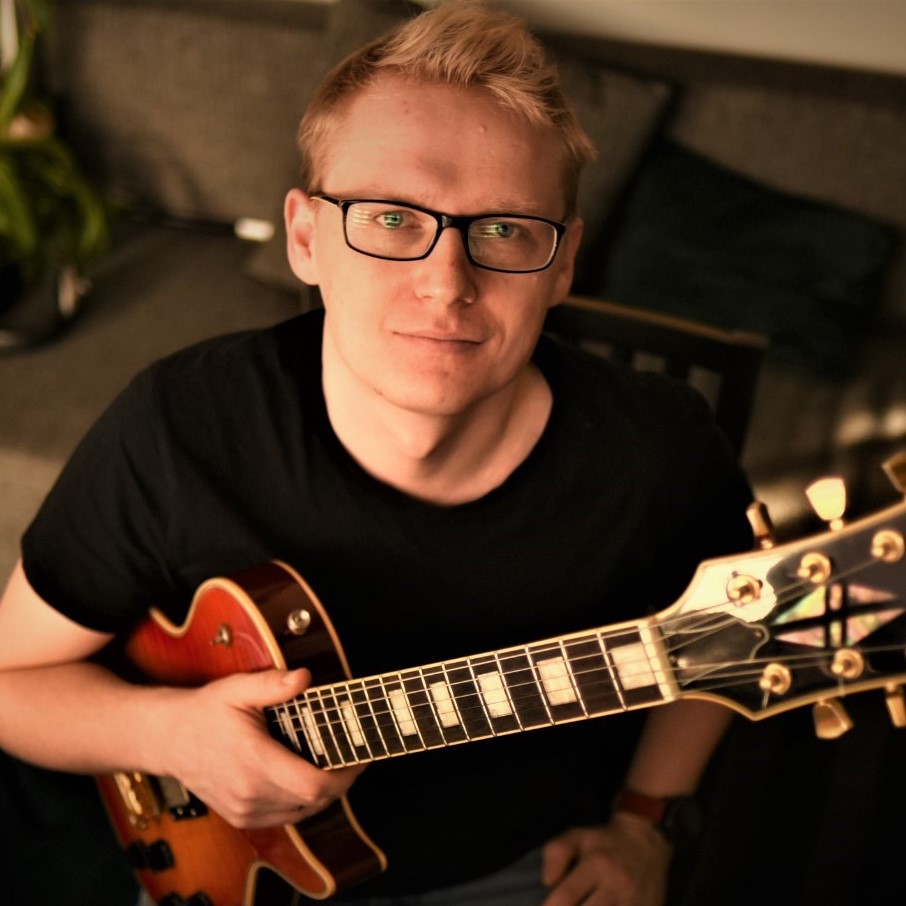 Gitarzysta Przemysław Hanaj gra na instrumencie, widoczne są struny i pudło rezonansowe gitary w kolorze brązowym, artysta nosi okulary i ubrany jest w czarną koszulkę