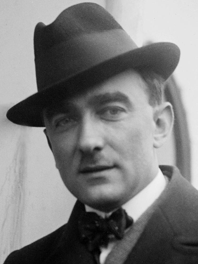 Kompozytor Karol Szymanowski jest mężczyzną w średnim wieku, ma okrągła twarz, ubrany jest w garnutur, a na głowie nowi kapelusz