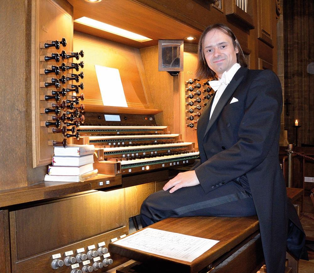 Organista Tomasz Orlow siedzi przed klawiaturą organów, jest ubrany w czarny smoking i białą koszulę z białą muszką