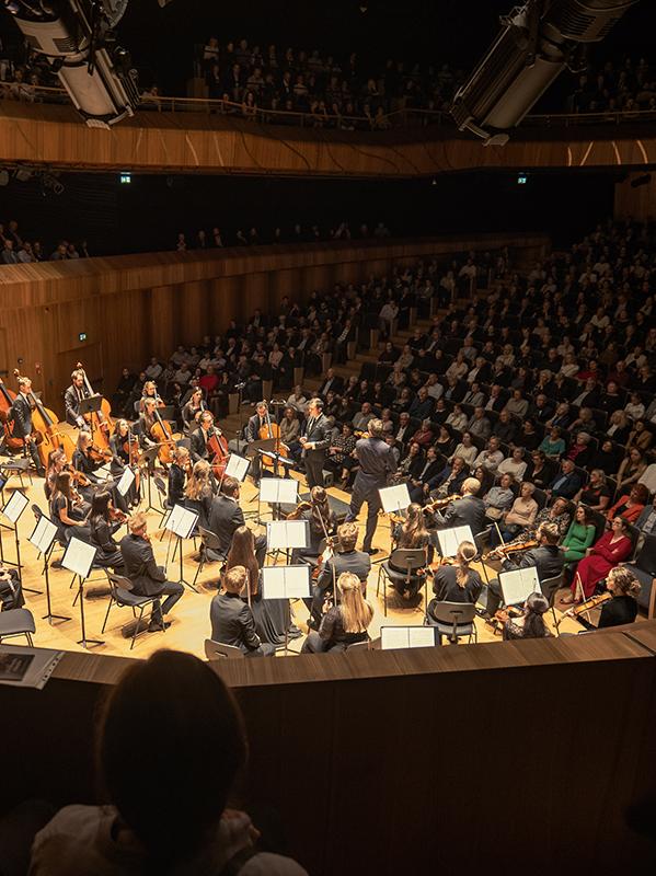 Muzycy orkiestry siedzą na mocno oświetlonej estradzie, trzymają w rękach różne instrumenty